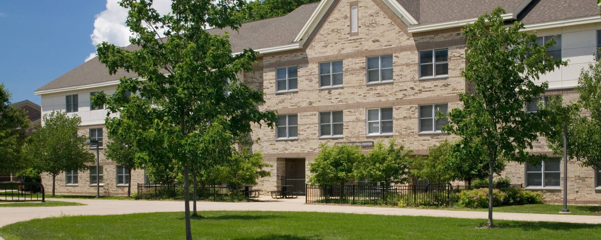 A three-story brick building, Kirkpatrick Living Center, with white trim is surrounded by lush green trees and grass under a clear blue sky, conveying a serene, welcoming atmosphere.